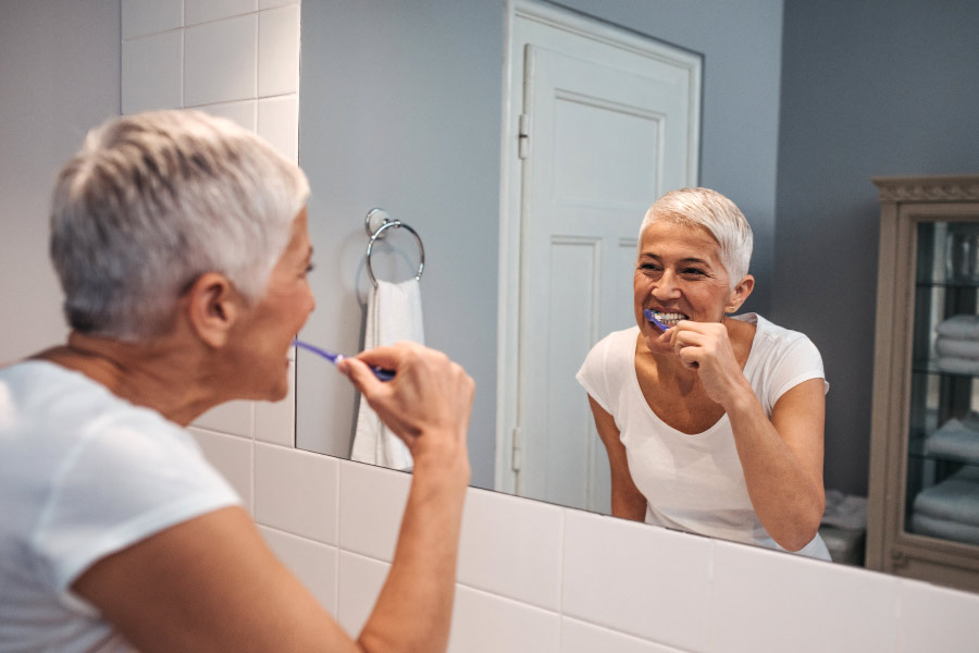 Lady with short gray hair brushing her teeth while looking in the bathroom mirror.