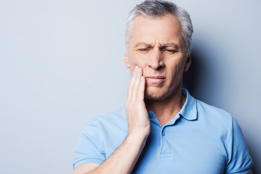 Gray haired man in a blue polo puts his hand to his cheek because of tooth sensitivity.