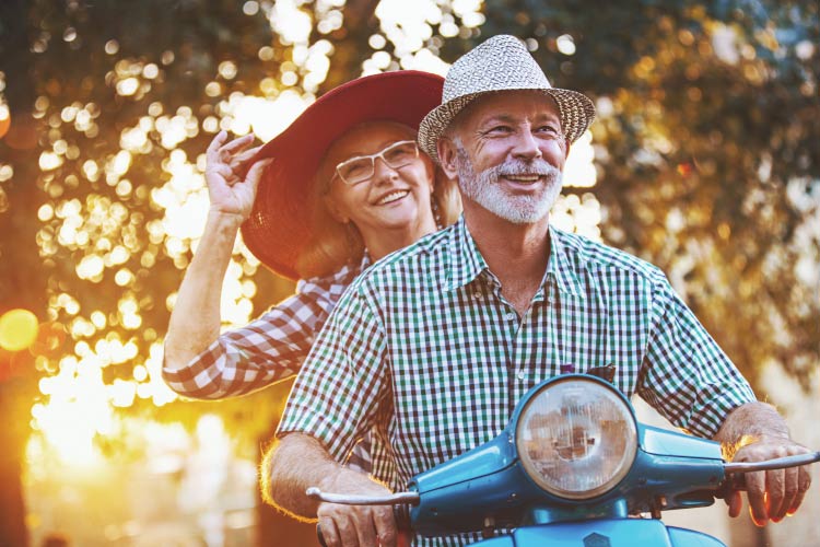 Woman with a big hat rides behind a man on a scooter with the sun filtering through the trees.